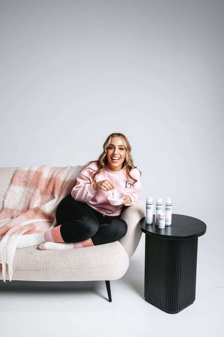 Person sitting on a couch with a blanket, holding a mug, and surrounded by bottles on a small table at TGS Beauty & Skin Perth