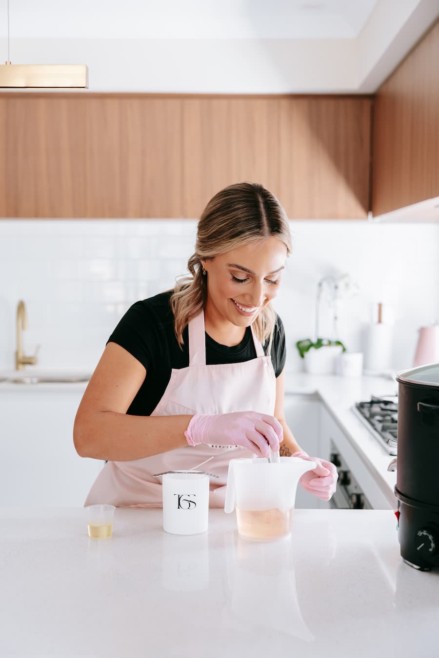 Woman in a kitchen preparing a drink, wearing an apron and gloves  at TGS Beauty & Skin salon in Perth.
