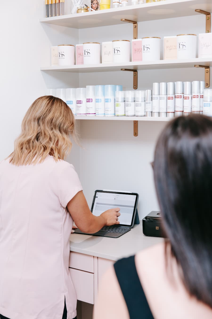 Two women in a store with shelves displaying various products and a woman using a laptop  at TGS Beauty & Skin salon in Perth
